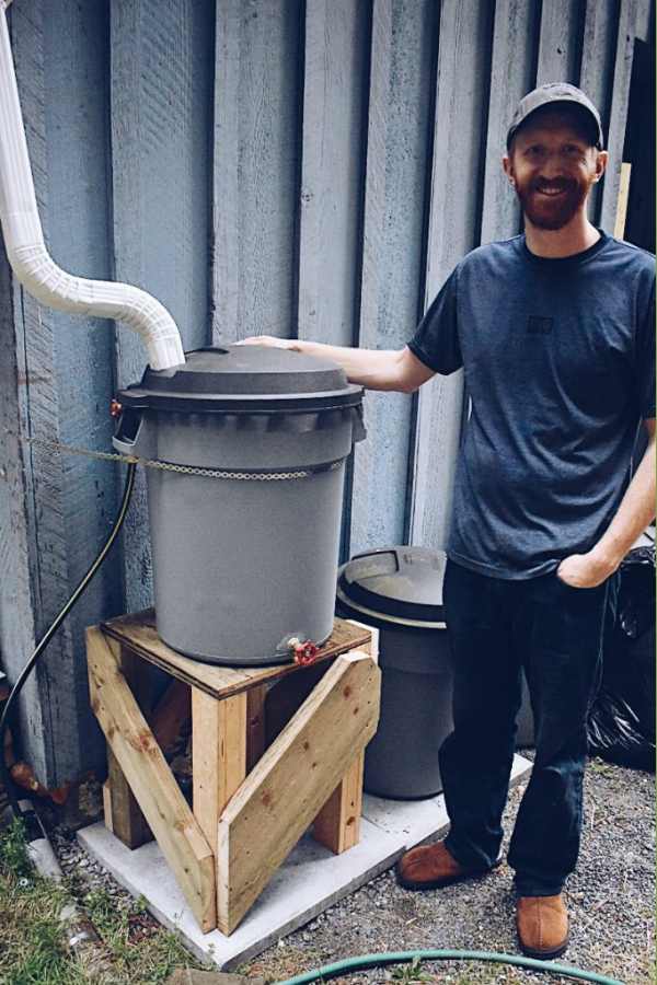 DIY wooden rain barrel stand by The Home and the Homestead.  Photo of a homeowner standing next to a rain barrel and his DIY wooden rain barrel stand.
